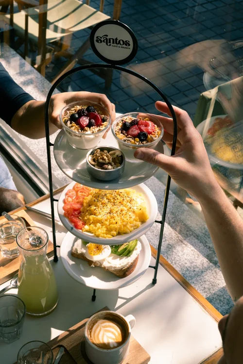 Plato de brunch Santos con huevo poché, tomates cherry, palta, frutos secos y pan integral junto a un café con leche